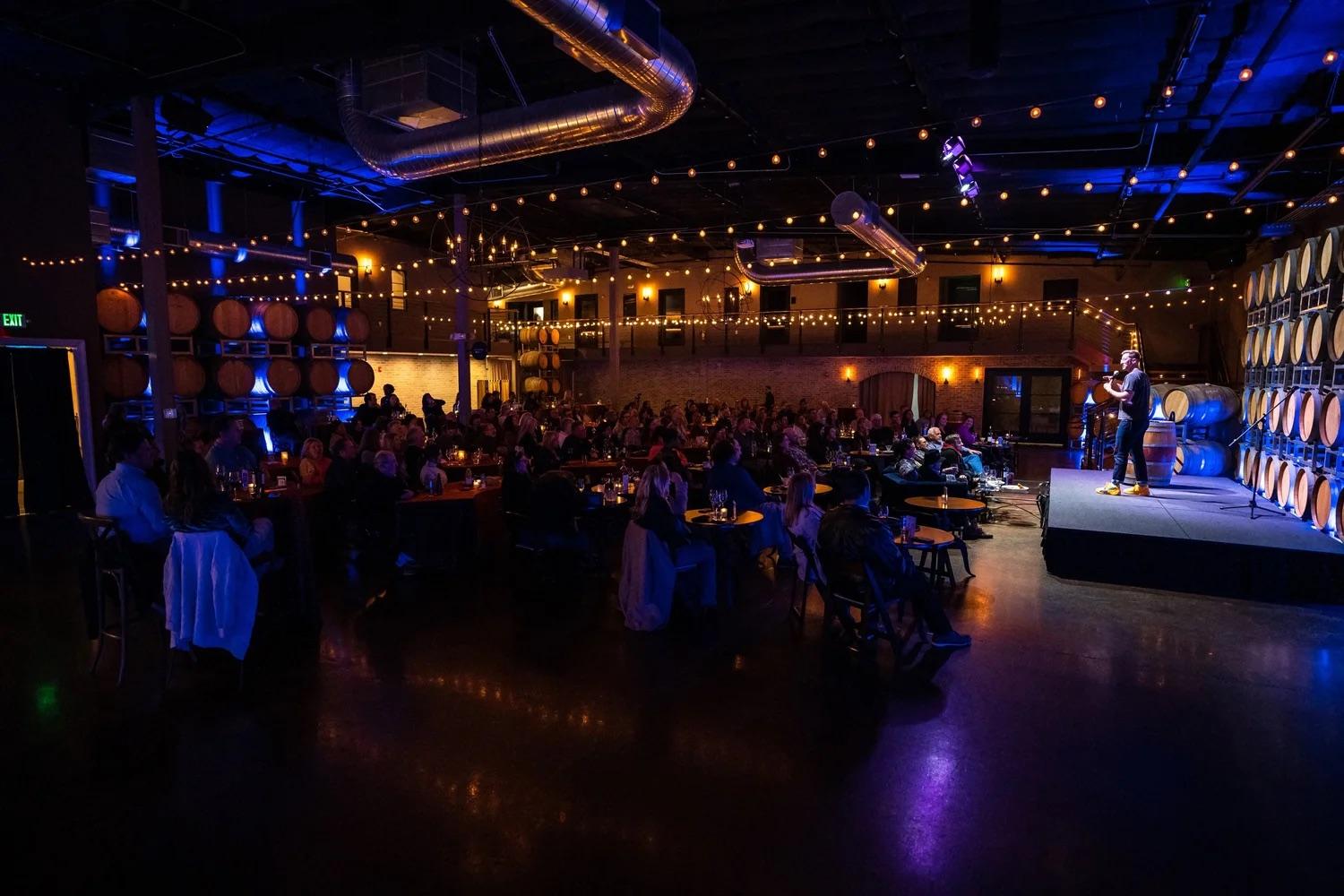 A full room of tables in Domenico Winery's barrel room, watching a comedian on a lit stage